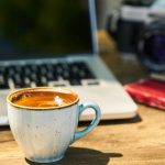 A cozy workspace with a cappuccino, laptop, and vintage camera on a wooden desk.